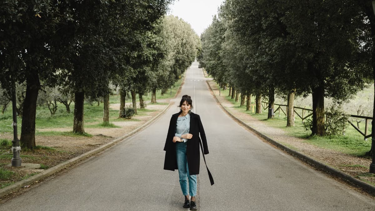 A fashionable woman stands confidently on a picturesque tree-lined road in Tuscany, Italy.